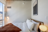 Sunlit bedroom with modern decor, featuring a comfortable bed with crisp white linens, accented with a warm orange throw blanket. Large window provides a city view, enhancing the serene ambiance.