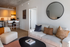Modern apartment living room featuring a cozy beige sofa with stylish cushions, a round wall mirror, and a chic open-plan kitchen with bar seating.