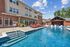Inviting apartment complex with luxurious outdoor pool area, elegant red lounge chairs, and charming brick architecture under a sunny sky.