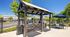 Charming outdoor grilling area with stainless steel grills under a rustic pavilion, perfect for entertaining. Adjacent modern seating with umbrella, surrounded by lush greenery and sunshine.