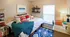 Cozy bedroom featuring a stylish bed with vibrant pillows, a soft blue throw, modern decor on shelves, and a functional study desk. Bright window light enhances the inviting atmosphere.