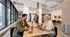 Modern co-working space with natural light, featuring people working and socializing over coffee at a large wooden table.
