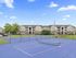 Spacious apartment complex with a vibrant blue tennis court and lush green surroundings under a clear blue sky.