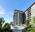 Modern apartment complex with elegant balconies overlooking a pristine swimming pool, surrounded by lush greenery and clear blue skies.