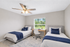 Bright and inviting bedroom featuring twin beds with elegant blue accents, a wooden desk by a large window, and a ceiling fan for comfort.
