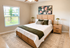 Bright and inviting bedroom with elegant wooden furniture, featuring a cozy bed adorned with stylish pillows and a green throw, complemented by natural light from a large window and vibrant floral artwork.