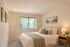 Serene bedroom with cozy bed, plush pillows, elegant artwork, and warm natural lighting through large window blinds.