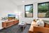 Bright and inviting living room with modern decor, featuring a sleek TV stand, stylish glass coffee table, and a cozy tan sofa accented by plush cushions. Large windows offer a tranquil view of greenery, enhancing the spacious feel.
