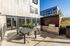Modern apartment patio with sleek furniture, outdoor grill, and large windows, set against a sunny blue sky backdrop.