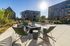 Charming outdoor patio with cozy seating around a modern firepit, surrounded by lush landscaping and stylish apartment buildings under a bright blue sky.