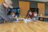 Joyful family creativity time in a bright, modern space with a father and mother helping young children color at a wooden table, surrounded by colorful art supplies.