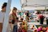Vibrant outdoor market with smiling visitors enjoying fresh flowers and coffee under sunny skies.