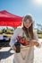 Cheerful woman at a vibrant outdoor market holding a fresh bouquet of flowers and a to-go coffee cup, standing under a bold red canopy with a bright sky overhead.