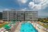Contemporary apartment complex with a vibrant pool area, surrounded by sun loungers and green umbrellas, overlooking stunning waterfront views under a bright blue sky.