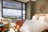 Inviting bedroom with plush bedding, wicker chair, and expansive window offering stunning urban views.