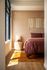 Sunlit bedroom with modern decor, featuring a cozy bed with rich burgundy bedding, elegant wooden floors, and a stylish woven wall hanging.