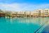 Luxurious apartment complex featuring a sparkling blue swimming pool, modern lounging areas, and stunning architecture under a clear blue sky.