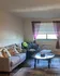 Cozy living room with a stylish gray sofa adorned with floral pillows, a large window with purple drapes, and a plush chair. Inviting atmosphere with elegant decor.