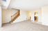 Bright and airy living space featuring a cozy carpeted area with elegant wooden stairs leading up. Modern kitchen with sleek appliances visible through an open doorway.