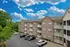 Charming three-story apartment building with brick fa&ccedil;ade, featuring spacious balconies and surrounded by lush greenery under a vibrant blue sky.
