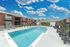Inviting apartment complex with a sparkling outdoor pool, sun loungers, and charming red brick buildings under a vibrant blue sky with fluffy clouds.