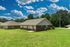 Charming single-story brick home with a spacious green lawn and picturesque countryside backdrop under a vibrant blue sky.