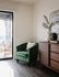 Elegant sunlit corner with a luxurious green armchair, stylish wood dresser, and balcony view, perfect for relaxation and enjoying serene evenings.