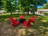 Charming outdoor area with vibrant red Adirondack chairs around a cozy fire pit, surrounded by lush greenery and nearby apartment buildings.