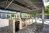 Luxurious outdoor kitchen with stainless steel appliances under a shaded pavilion, surrounded by lush greenery and poolside seating.