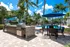 Luxurious outdoor space featuring a sparkling pool, modern barbecue station, and relaxing patio furniture under vibrant blue umbrellas, surrounded by lush palm trees.