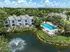 Arial view of elegant townhouses adjacent to a shimmering swimming pool and serene pond with a fountain, surrounded by lush greenery under a bright blue sky.