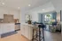 Modern open-concept kitchen and living room with sleek white cabinetry, stylish island seating, and a view of lush greenery through large glass doors.