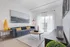 Bright and modern living room featuring a plush blue sofa adorned with yellow accent pillows, a sleek round coffee table, and a stylish lamp. Natural light pours through sheer curtains, highlighting vibrant decor pieces and city views from the balcony.