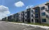Modern three-story apartment complex with private balconies, surrounded by lush greenery under a vibrant blue sky.
