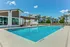 Luxurious apartment pool area with stylish pergola and seating, surrounded by lush landscaping and modern buildings under a bright blue sky.