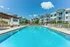 Elegant apartment complex with modern white facades featuring a sparkling swimming pool, comfortable lounge chairs, and a relaxing pergola under a clear blue sky.