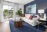Chic living room with a bright, airy vibe featuring a white sofa, abstract art, and a glass coffee table against a deep blue accent wall. Sliding doors open to a lush outdoor view.