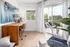Bright home office space with a modern wooden desk, elegant decor, large abstract painting, and a serene view through sliding glass doors leading to a balcony with lush greenery.