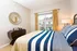 Inviting bedroom with stylish striped bedding, elegant dark wood dresser, and a modern circular mirror. Bright natural light streams through large windows with golden curtains, creating a warm and peaceful ambience.