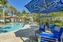 Luxurious poolside oasis with modern blue lounge seating and striped umbrellas, surrounded by lush palm trees and elegant villas.