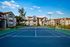 Full-sized community tennis court with a blue playing surface, surrounding fencing, and apartment homes in the background under a sunny sky.