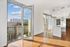 Modern apartment interior with large floor-to-ceiling glass doors open to a city view, featuring wood flooring and a sleek white kitchen island.