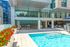 Contemporary outdoor pool area with turquoise water, surrounded by white tile flooring, red flower planters, a covered bar with high stools, and a shaded lounge seating area.