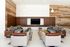 Modern lounge area featuring two sofas and four armchairs, a large flat-screen TV mounted on a wood-paneled wall, glass-top coffee tables, and stylish pendant lights.