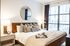 Cozy bedroom with a leather-frame bed, layered pillows, large windows with curtains, and a decorative round mirror.