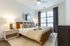 A cozy, modern bedroom featuring a leather-framed bed, neutral bedding, a circular mirror, and large windows with blackout curtains.