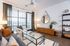 Stylish living room featuring a glass coffee table, gray sofa with orange and blue pillows, white accent chair, wooden console, round mirror, and large windows with sheer green curtains.