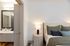 Partial view of a cozy bedroom with a tufted cream headboard, green and navy bedding, nightstand with a modern lamp, and a glimpse into a sleek bathroom with a large mirror and wood vanity.