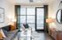 Modern living room with a gray couch, accent pillows, glass coffee table, and floor-to-ceiling windows with sheer green curtains.