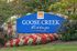 Blue entrance sign for Goose Creek Village, surrounded by neatly landscaped flowers and greenery, with a stone backdrop and trees in the background.